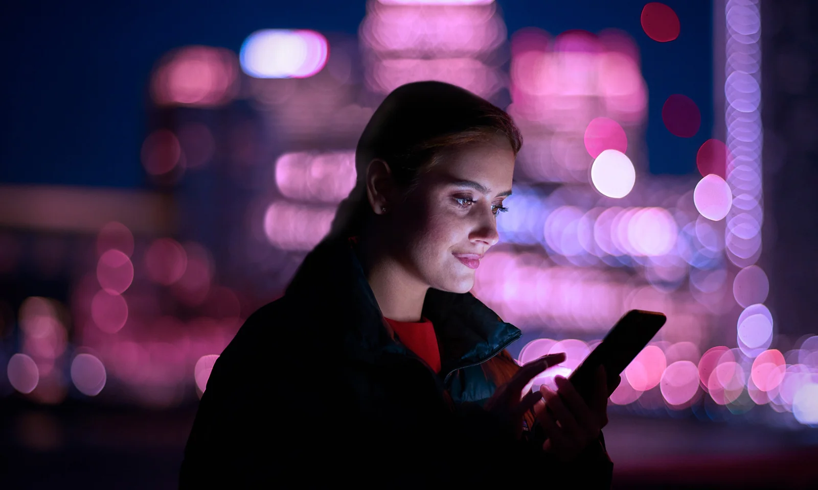 A woman using a smartphone against a backdrop of a cityscape illuminated by pink and purple lights, symbolising the transition of Guidewire to cloud-based solutions.