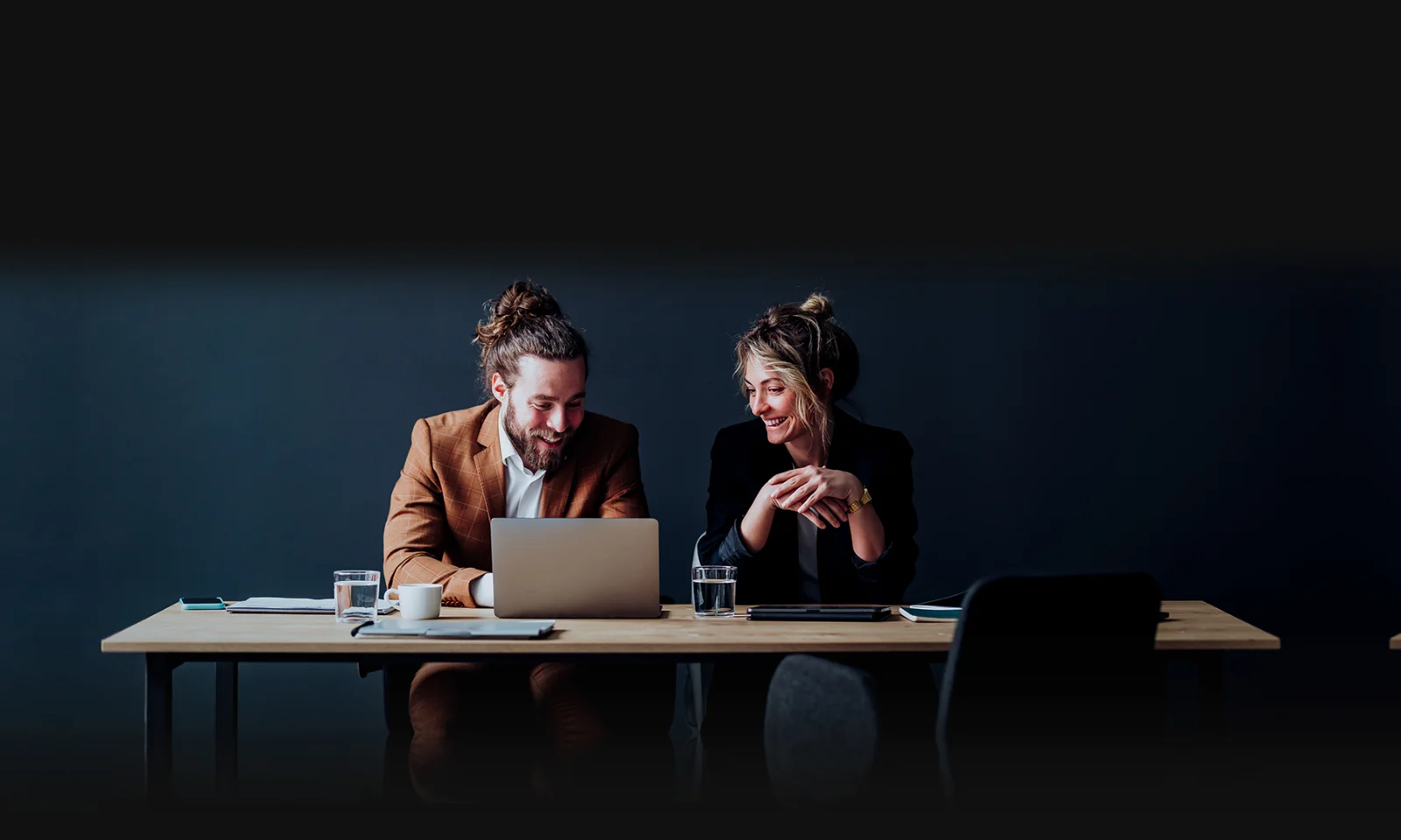 Two professionals, a man and a woman, engaged in a collaborative discussion while working on a laptop in a modern, minimalist workspace with a dark background.