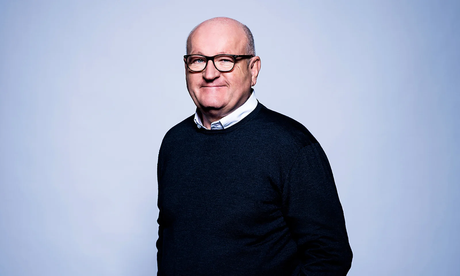 Ulrich Dietz posing with a smile against a blue background, wearing glasses and a dark sweater