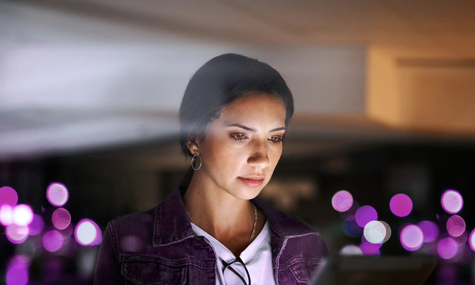 Focused woman using a digital device in a softly lit environment, with purple lights in the background.