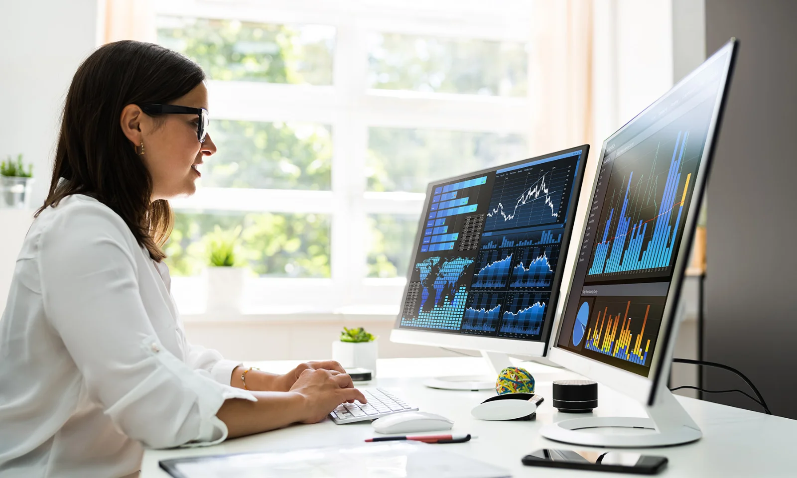 Woman in a white shirt working on a computer with dual monitors displaying various data charts and graphs in a bright office.