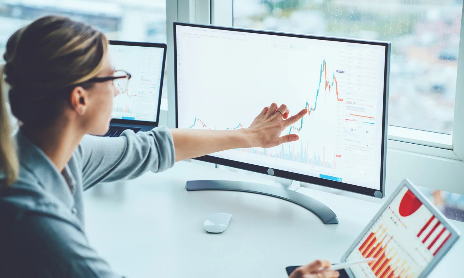 Businesswoman analyzing financial data on a large monitor, with charts and graphs displayed on multiple screens in a modern office.