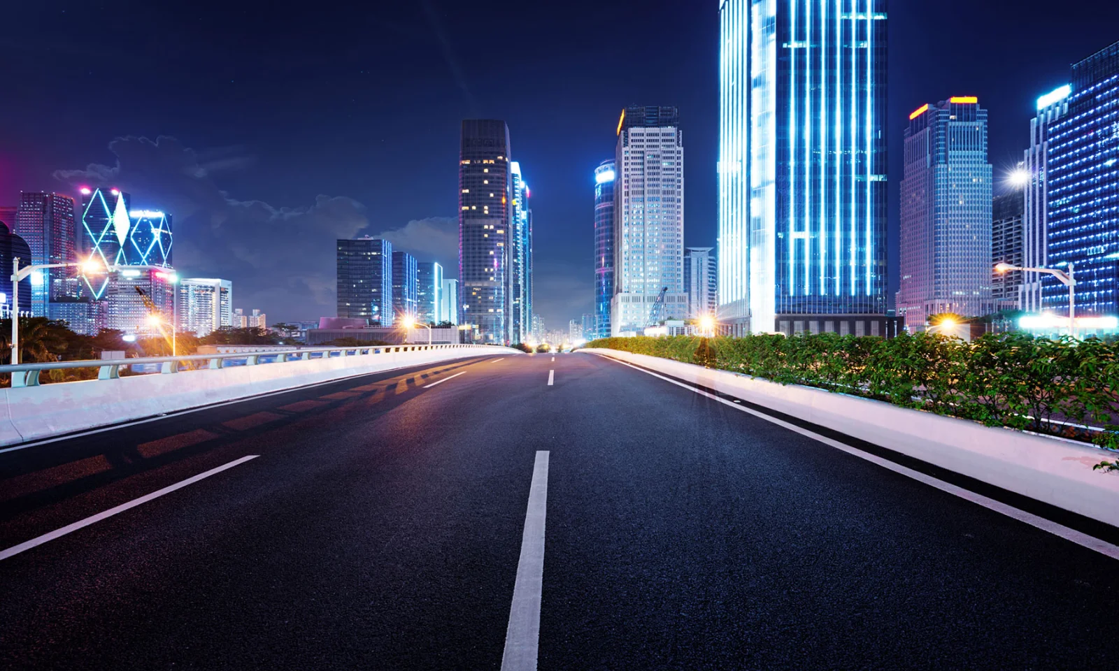 Illuminated cityscape at night with an empty highway leading towards skyscrapers.