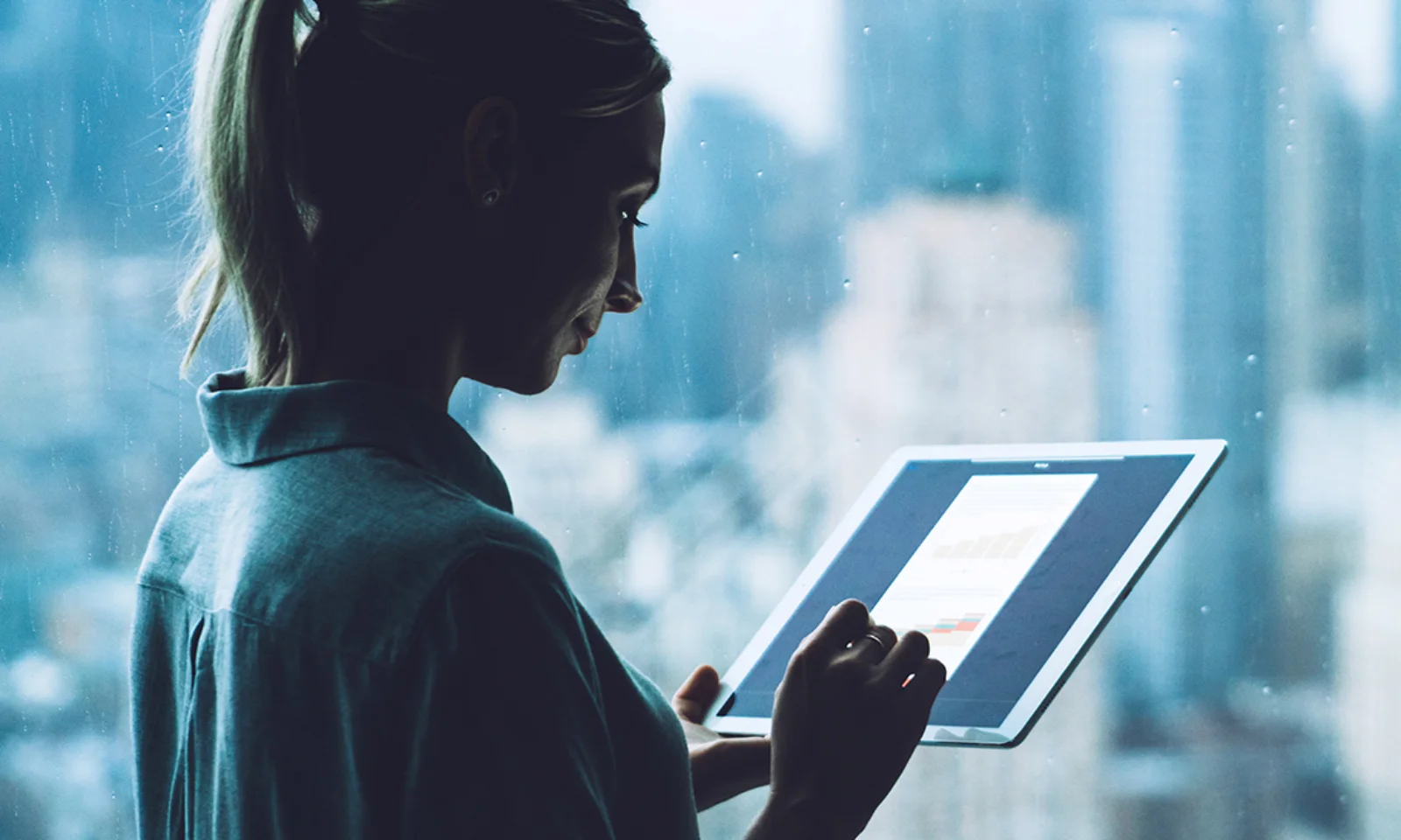 Silhouette of a professional woman analyzing data on a tablet while standing near a window overlooking a cityscape.