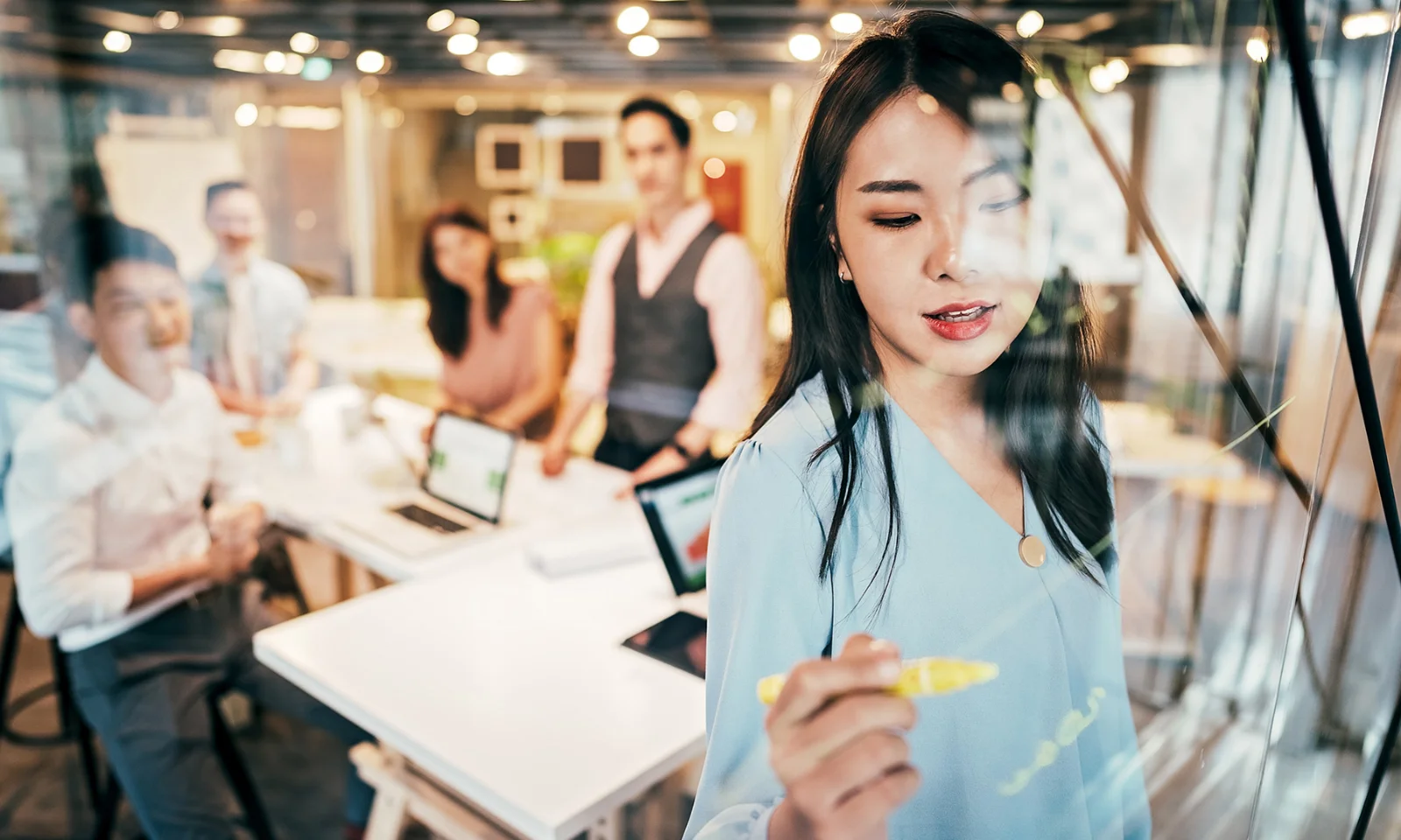 A woman writing on a glass board with a team in the background, representing GFT&#039;s culture and values.