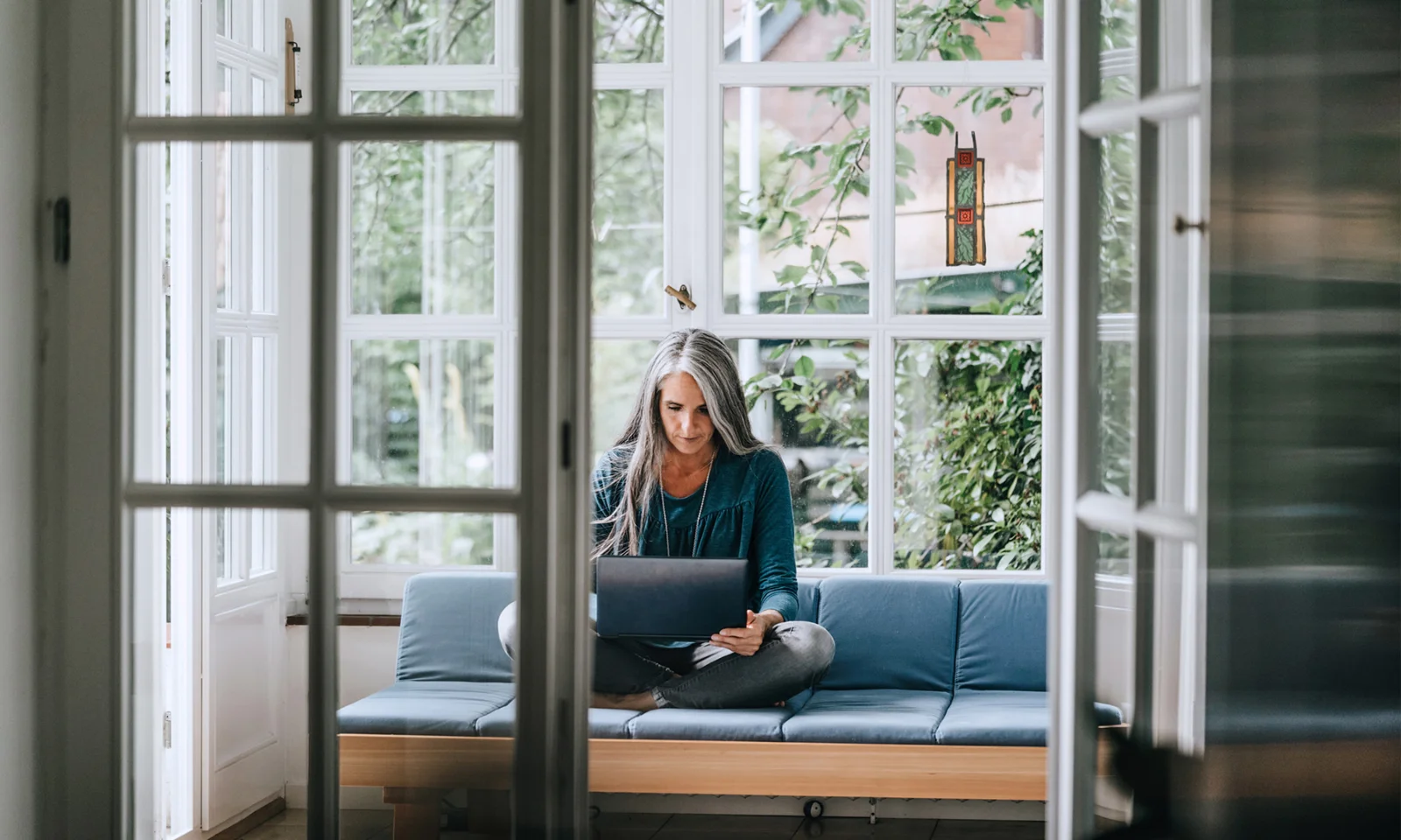 Woman working on a laptop while sitting on a couch in a bright room.