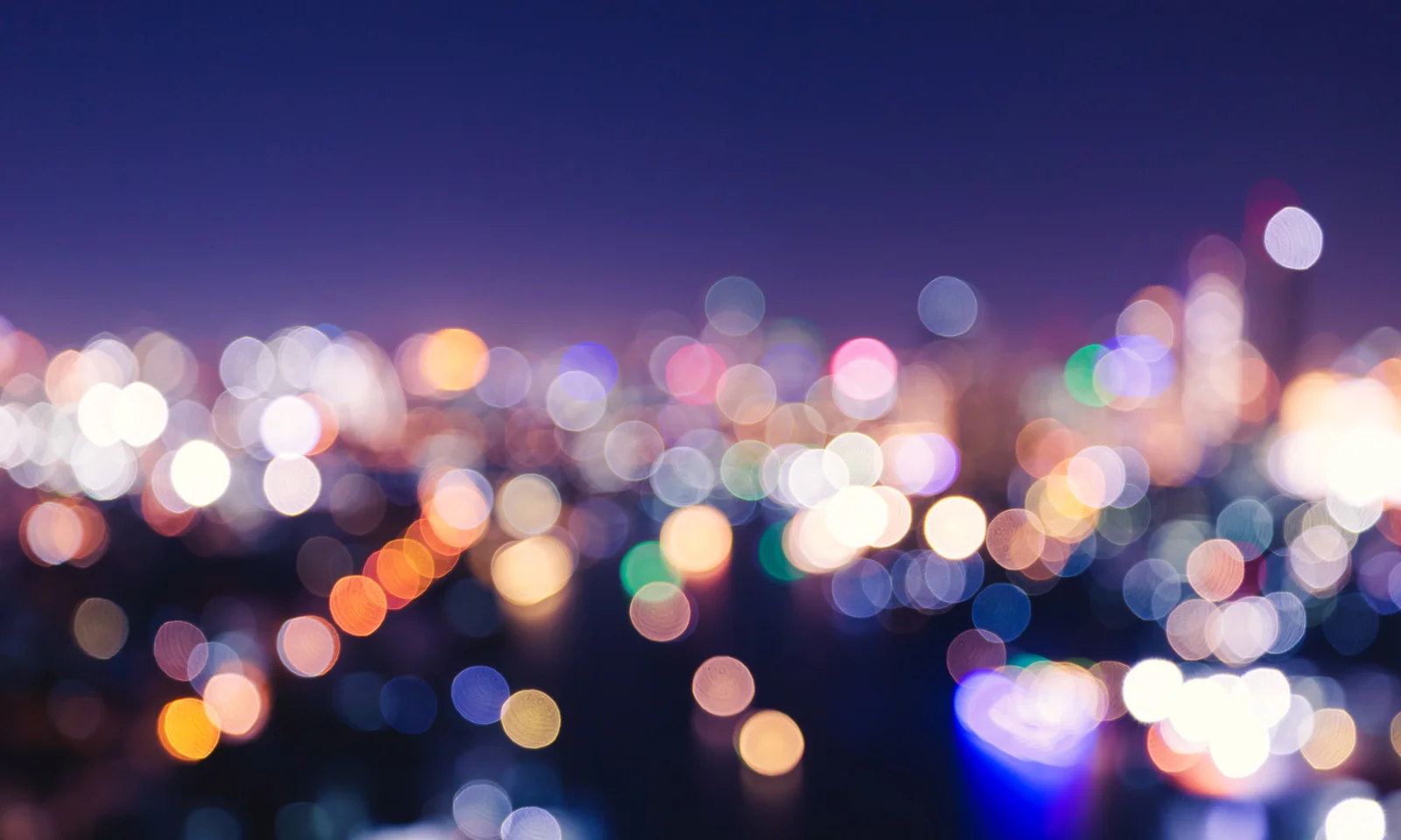 A blurred image of colorful city lights glowing against a dark night sky, creating a bokeh effect.