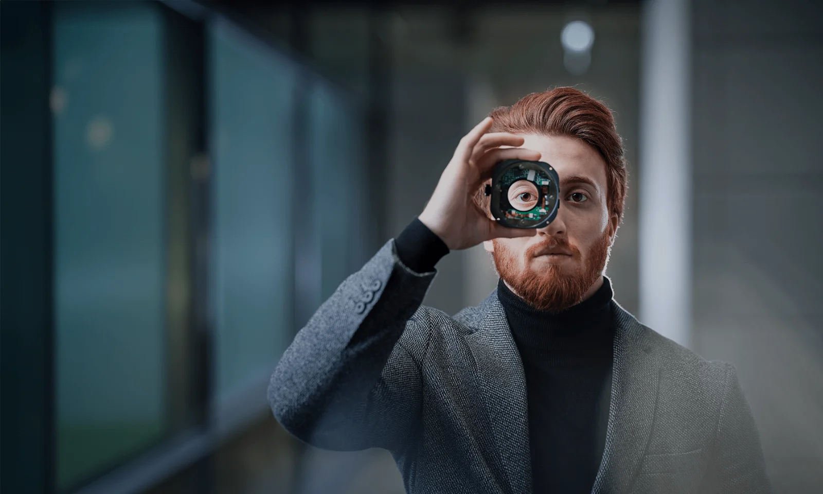 A man with red hair and beard holds a technological component in front of his eye, symbolizing innovation and focus. He is dressed in a grey suit and stands in a modern, dimly lit environment.