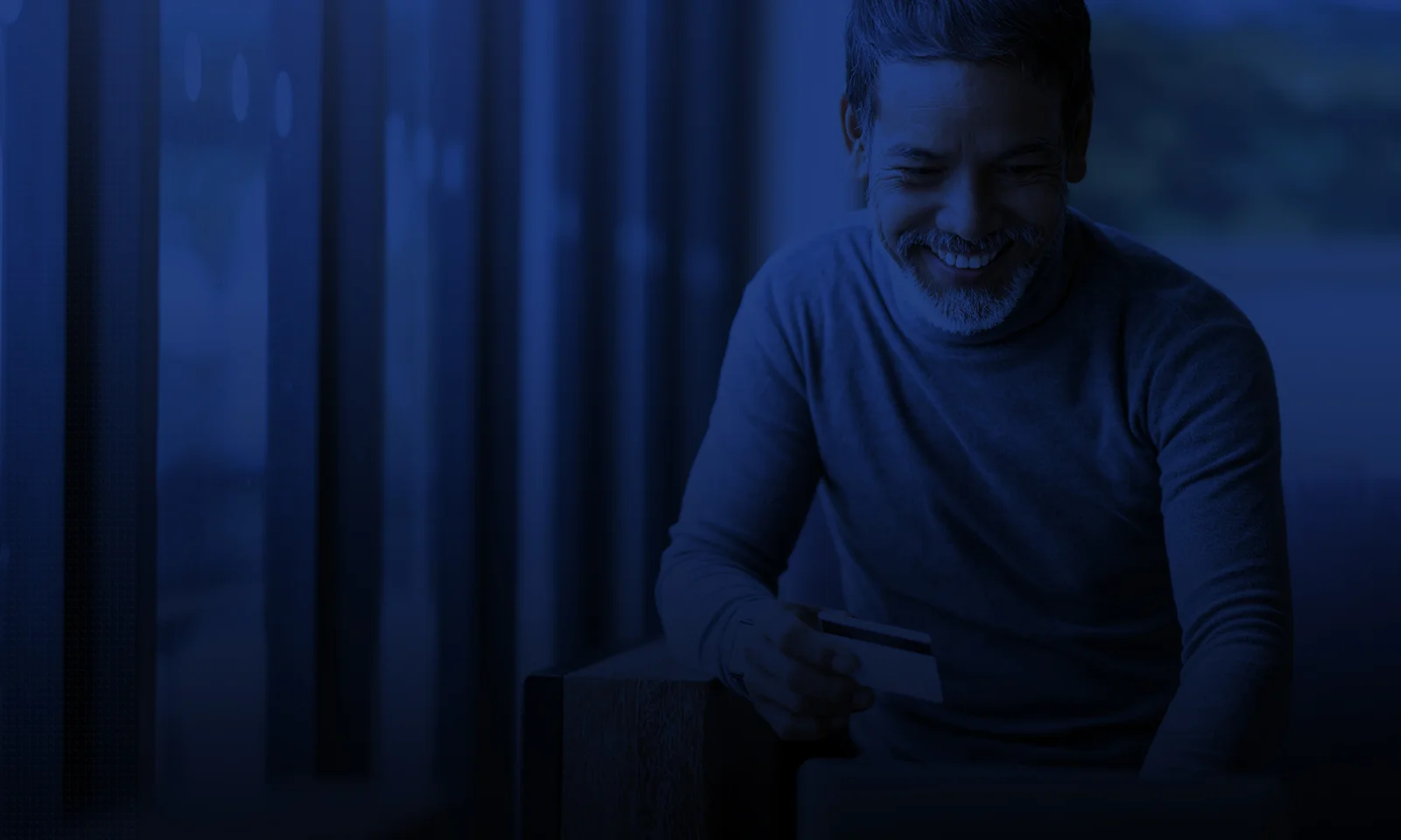 A man smiling while holding a credit card, making an online payment in a dimly lit, modern setting.