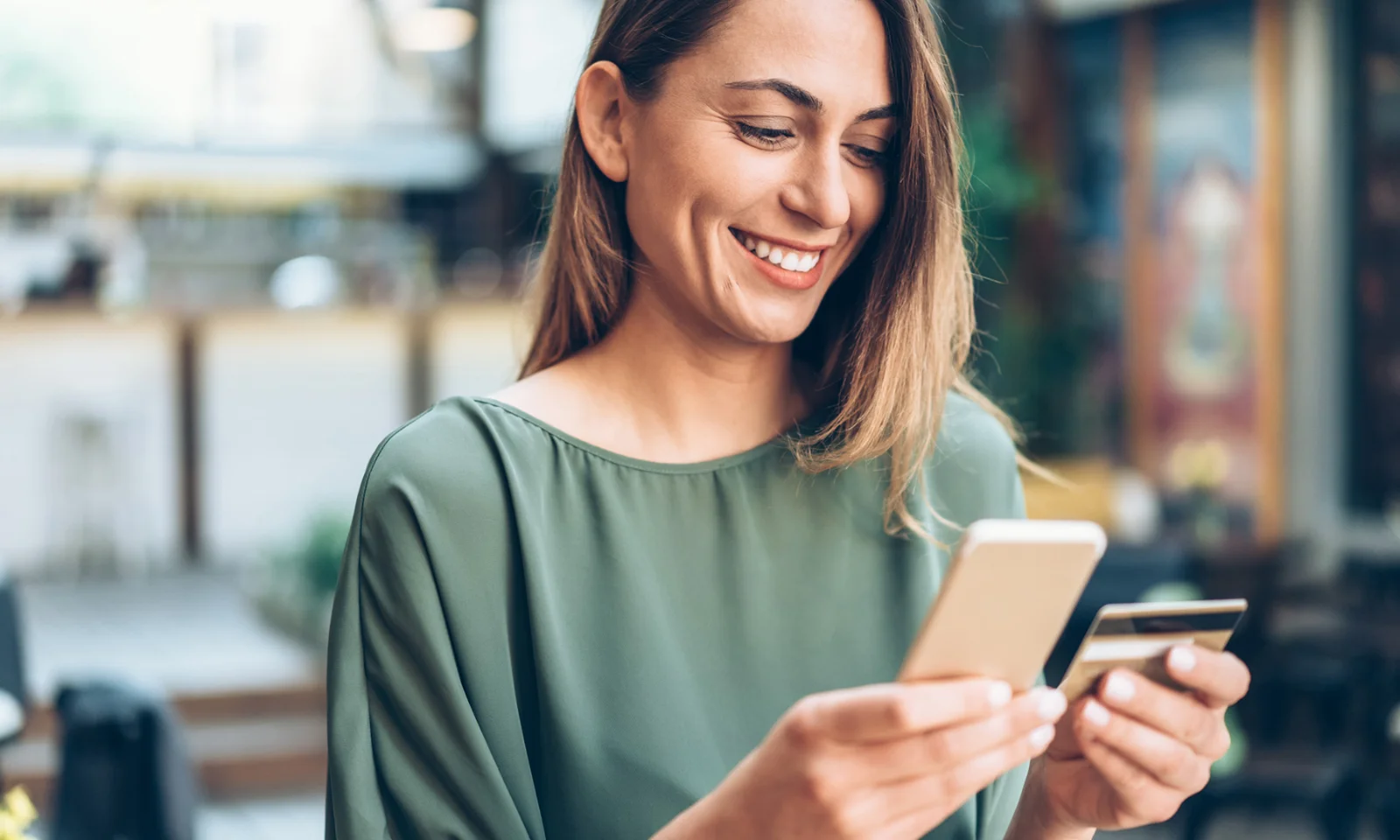 Una mujer sonriente usa un teléfono inteligente y sostiene una tarjeta de crédito, lo que representa la conveniencia de una nueva aplicación móvil de banca comercial.