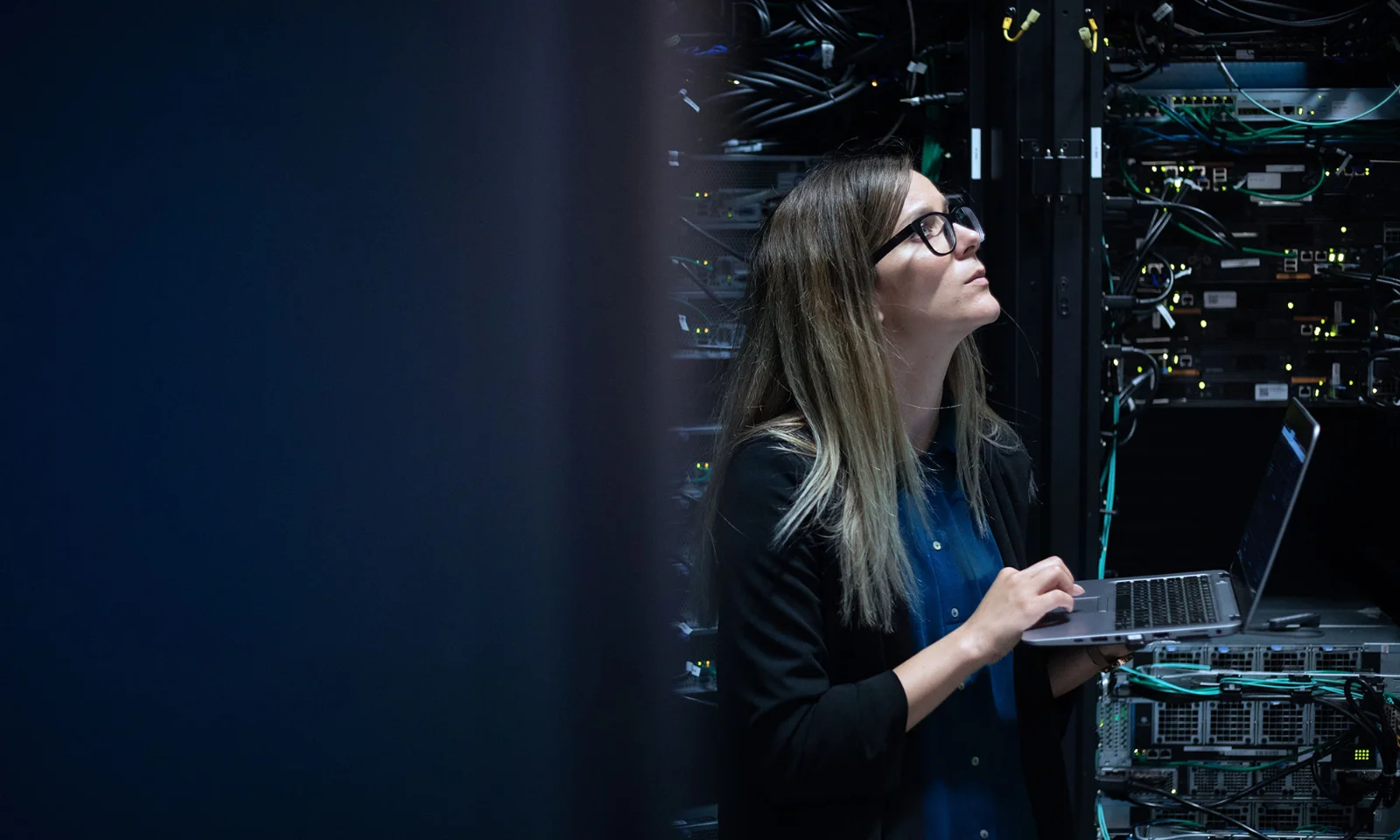 A female IT professional works on a laptop in a server room, surrounded by networking cables and server equipment, representing a high-performance computing (HPC) solution.