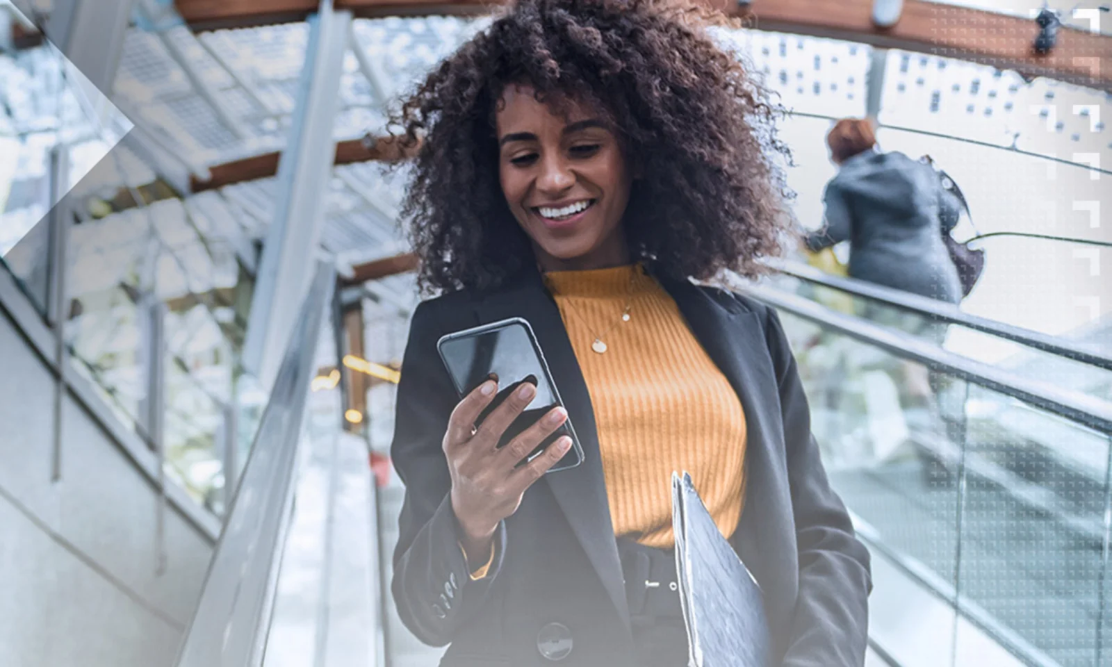 Smiling businesswoman using a smartphone while riding an escalator in a modern building with glass and metal architecture.