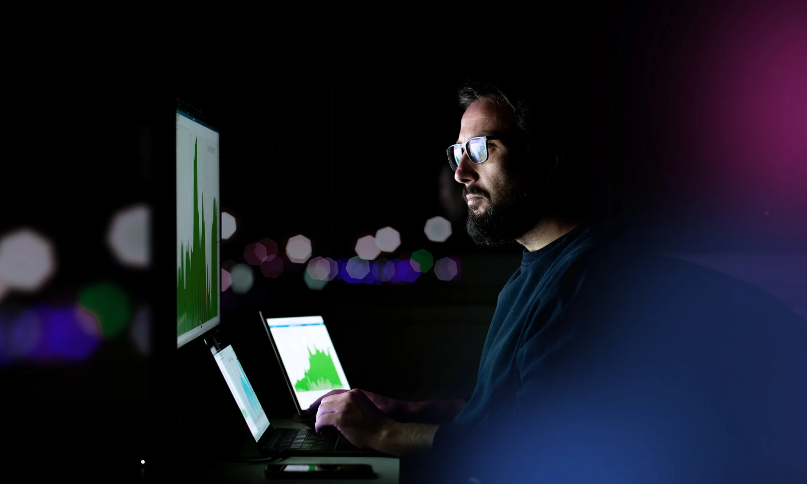 A man analyzing data on multiple screens in a dark room, focused on charts and graphs, with soft, blurred lights in the background.