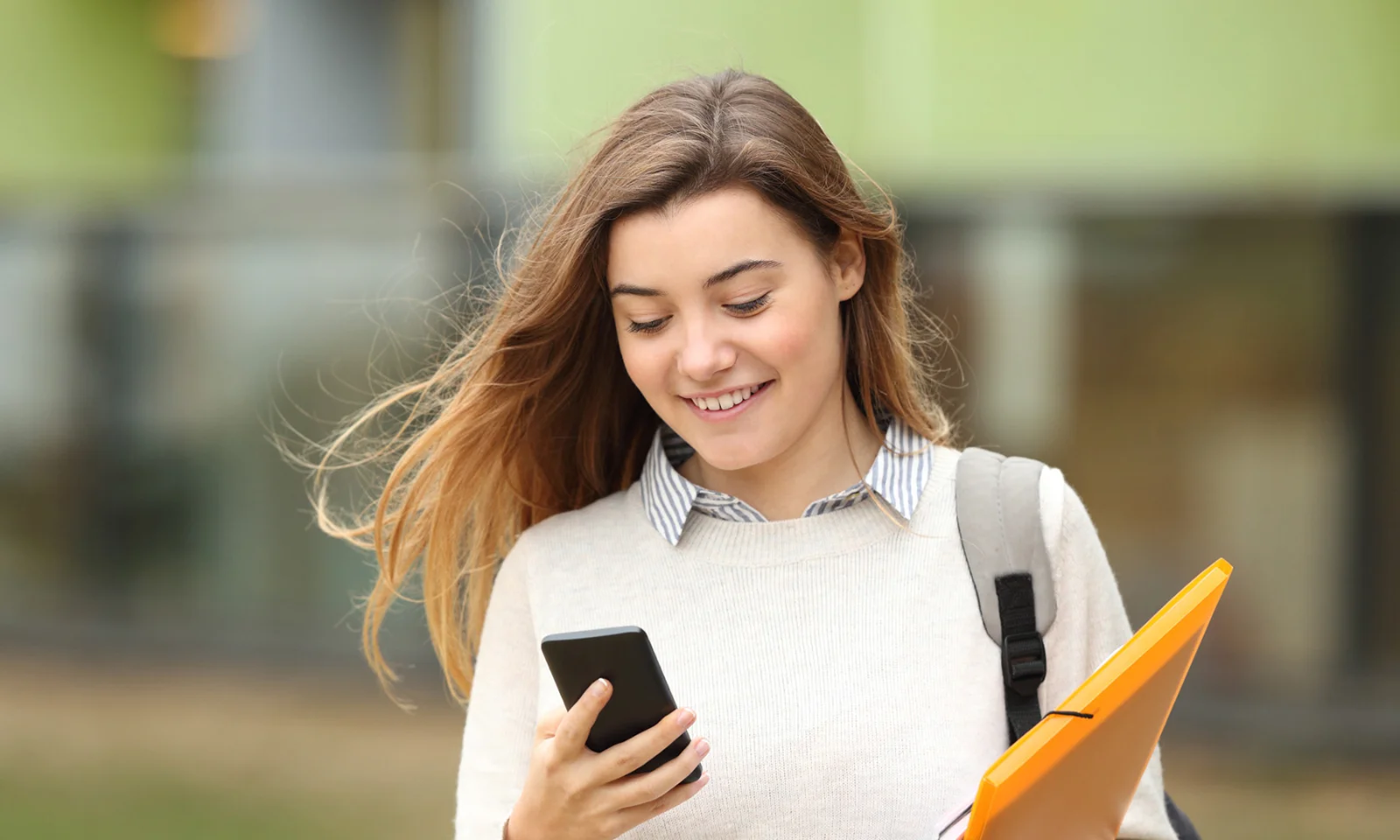 A young woman smiling while using her smartphone, holding a folder, symbolising the convenience and accessibility of the Crédit Agricole BankMeApp.