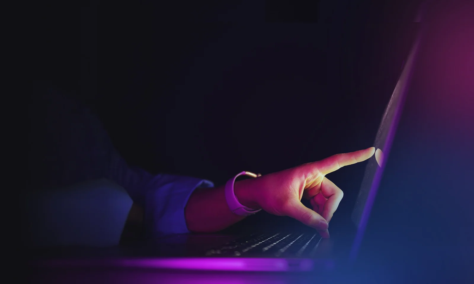 A hand pointing at a laptop screen in a dark setting, illuminated by purple and pink light, symbolising interaction and technology.