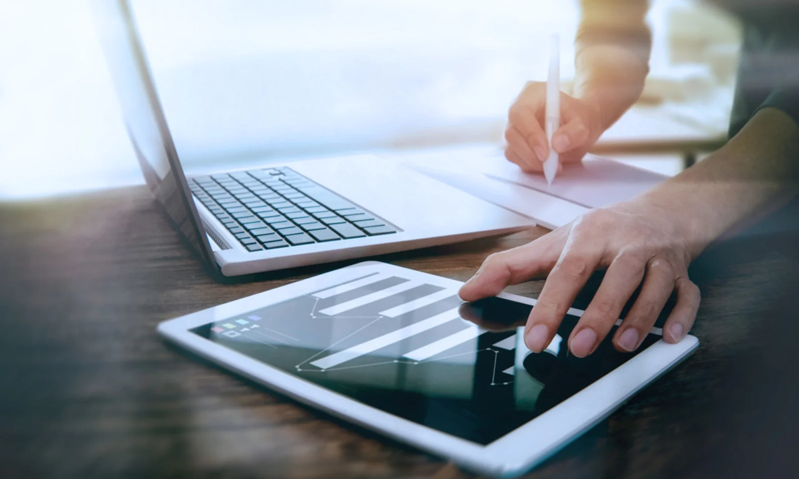 Close-up of a person working on a tablet with a laptop nearby, representing a modern digital work environment with multitasking and data analysis.