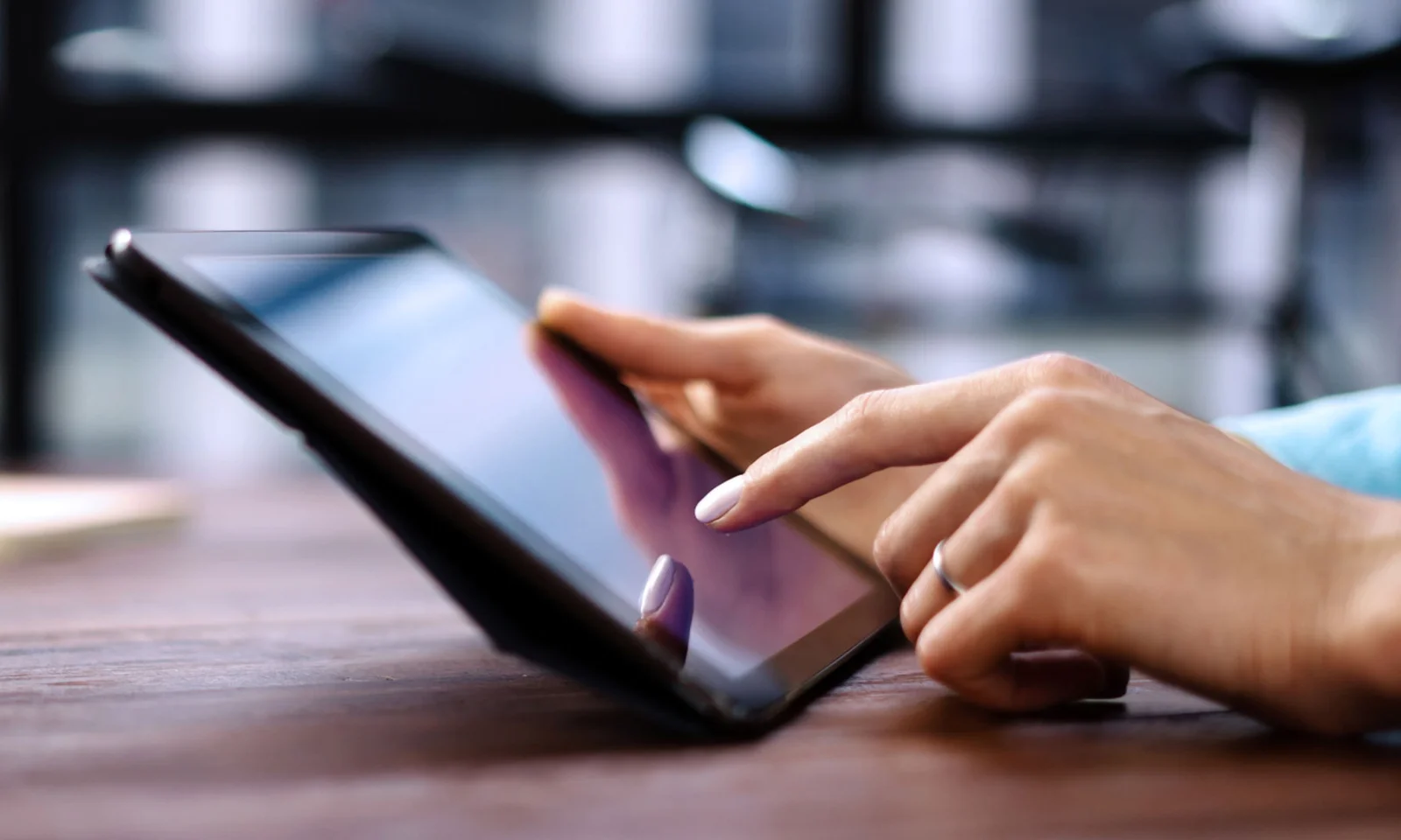 A imagem mostra um close-up de mãos usando um tablet, com uma mão tocando na tela. O tablet está sobre uma superfície e o fundo está levemente desfocado, sugerindo um espaço interior. O foco está na interação com o tablet.