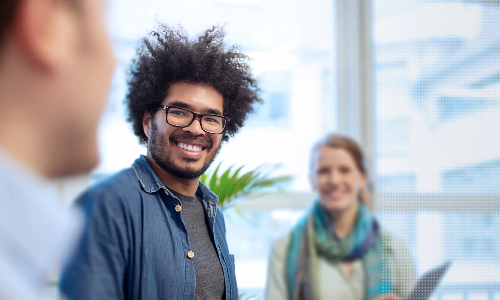 Smiling team members engaging in conversation in a bright office setting, with one man in the foreground wearing glasses and a denim shirt.