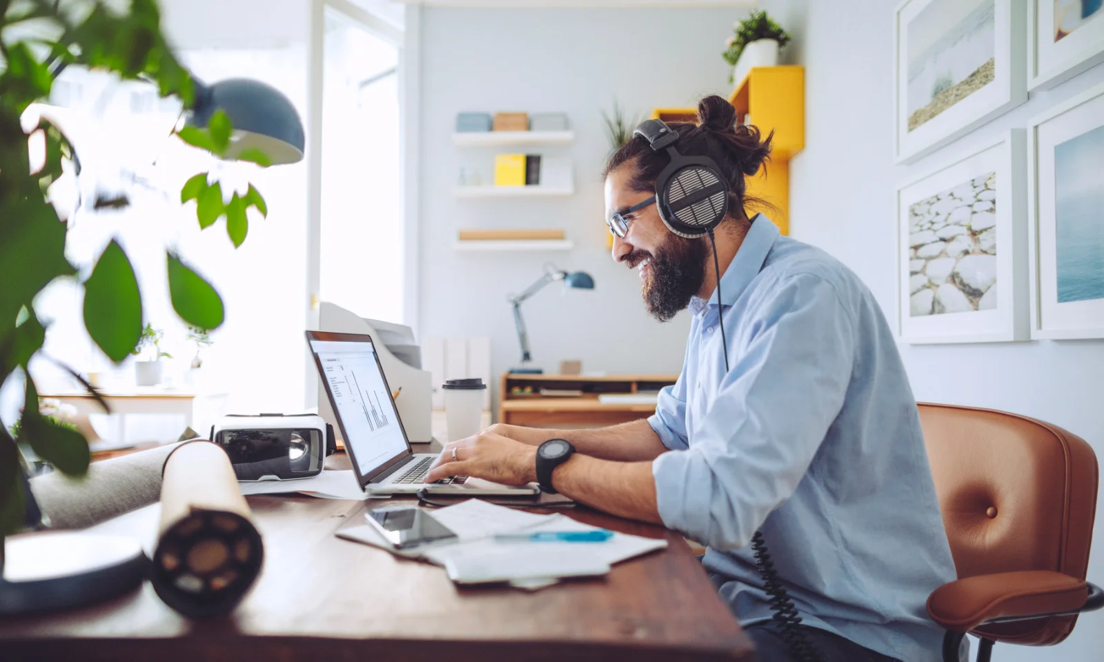 Man wearing headphones and glasses working on a laptop at home office, surrounded by tech gadgets and documents.