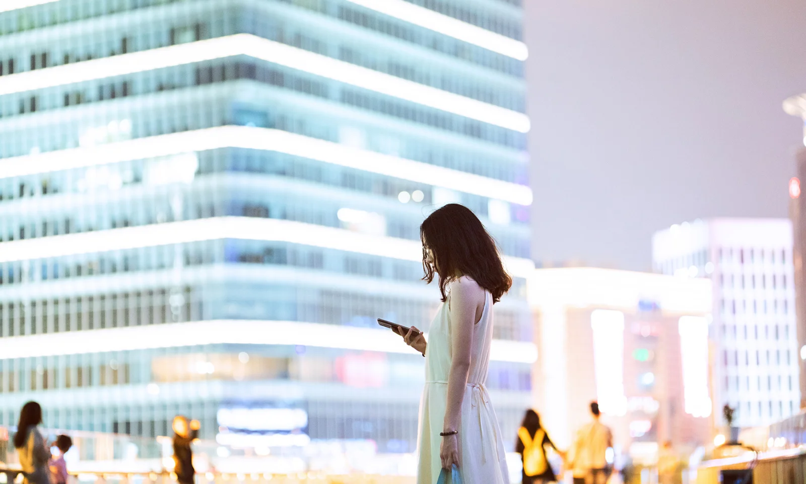 Uma mulher parada em um ambiente urbano à noite, olhando para seu smartphone, com prédios modernos bem iluminados ao fundo, representando o tema da digitalização no setor bancário.