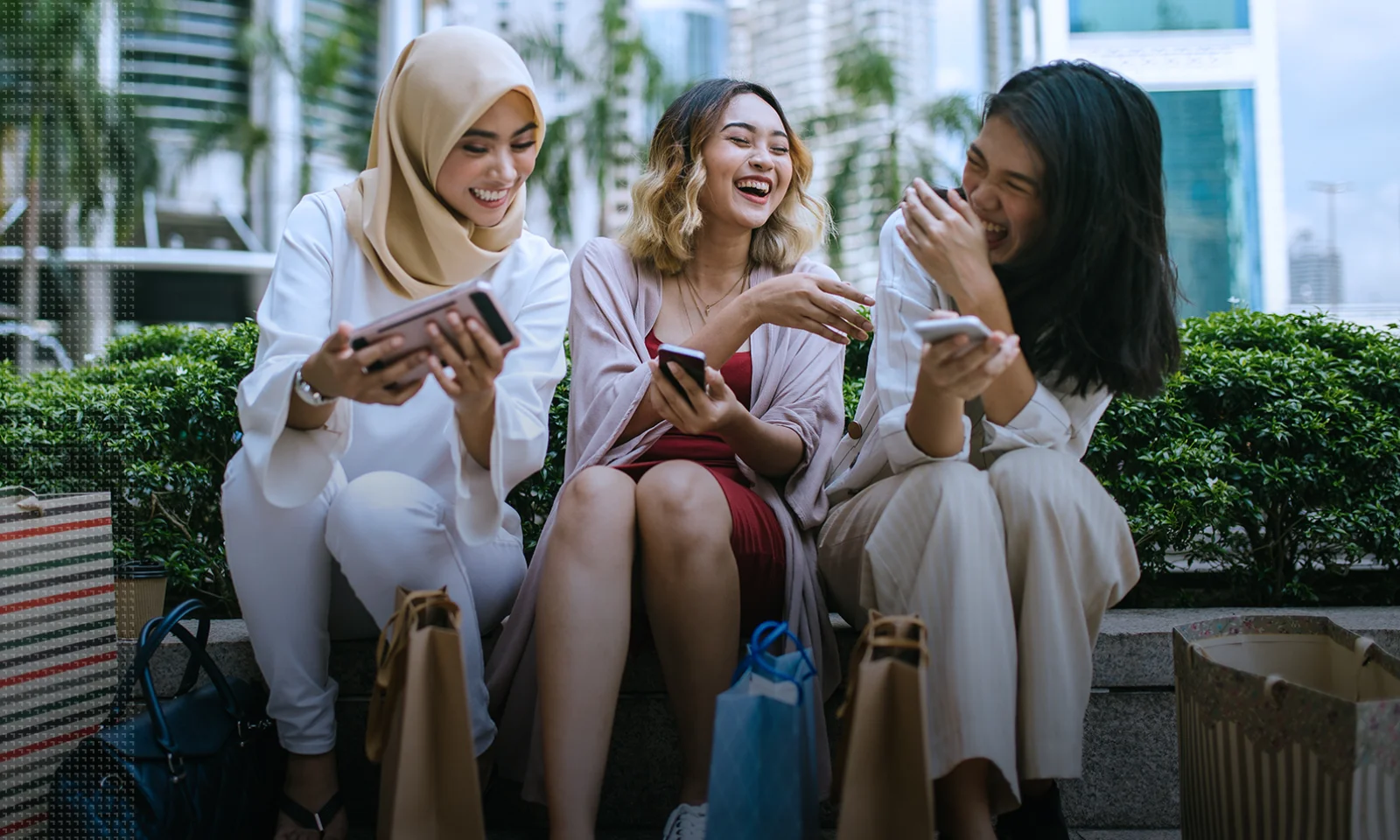 Three women sitting outdoors, laughing and sharing moments on their smartphones after a shopping trip, with shopping bags around them, symbolizing friendship and modern urban lifestyle.