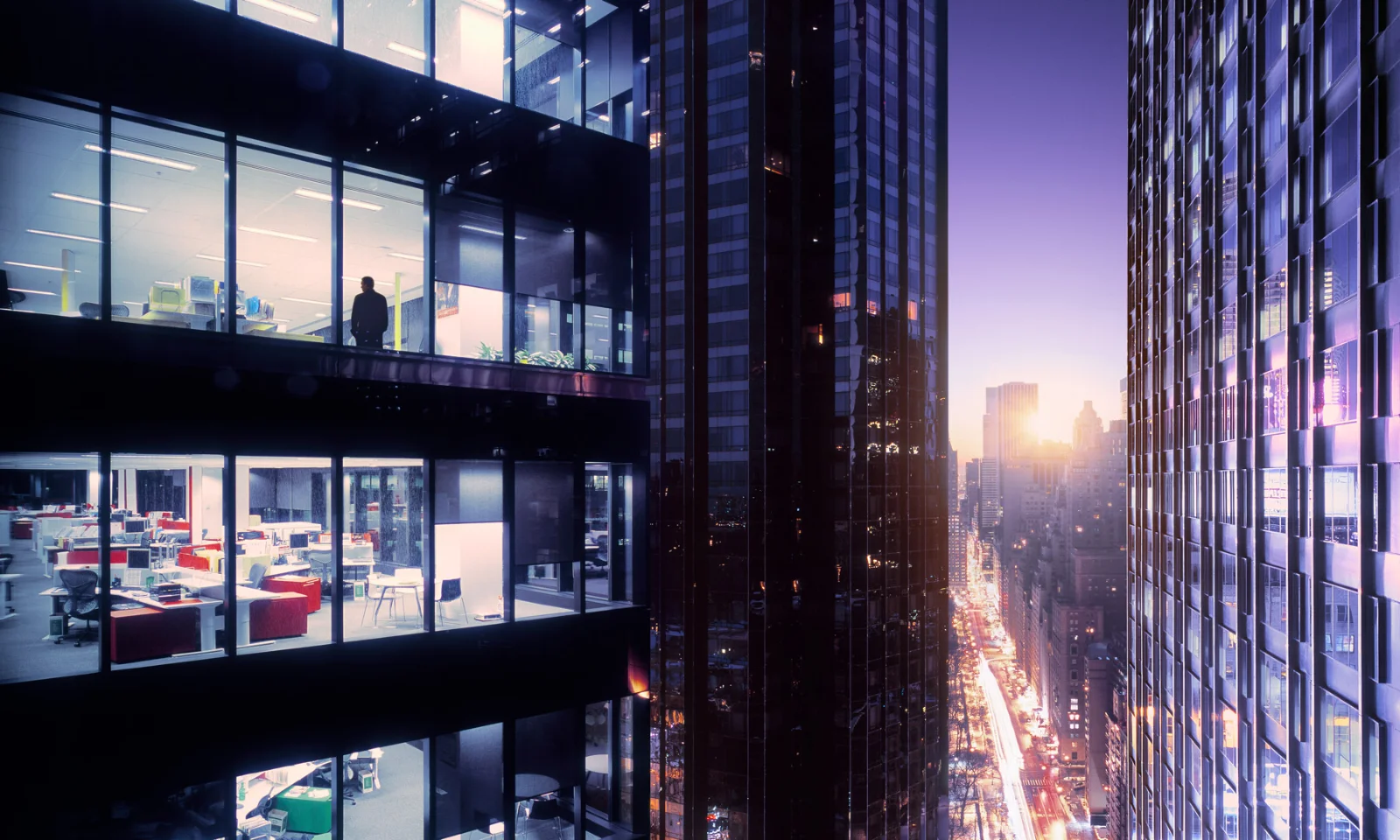 A cityscape at dusk with illuminated office buildings, one with a silhouette of a person inside, representing the evolving landscape of digital banking and financial services.