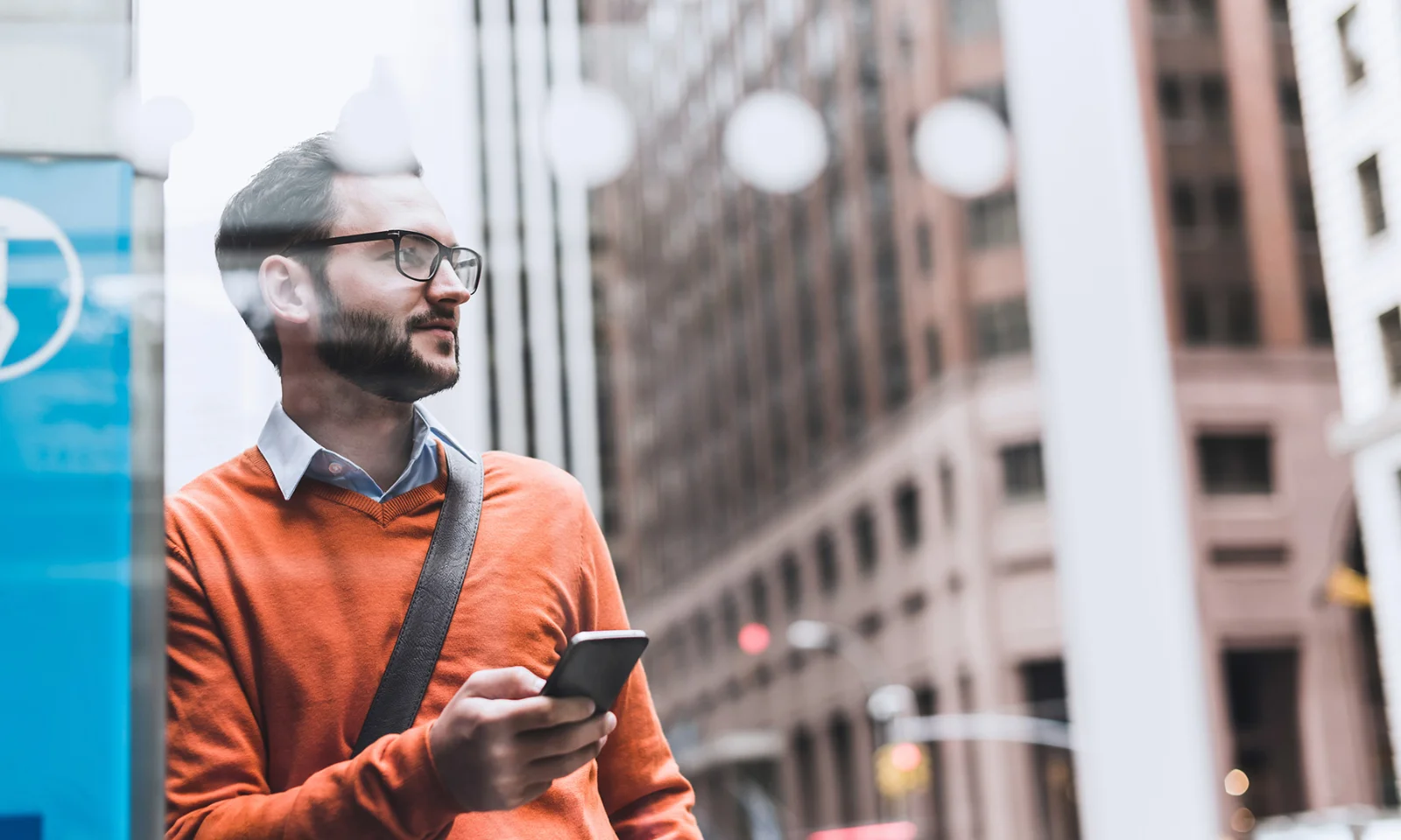 Man in an orange sweater holding a smartphone, standing in an urban environment.