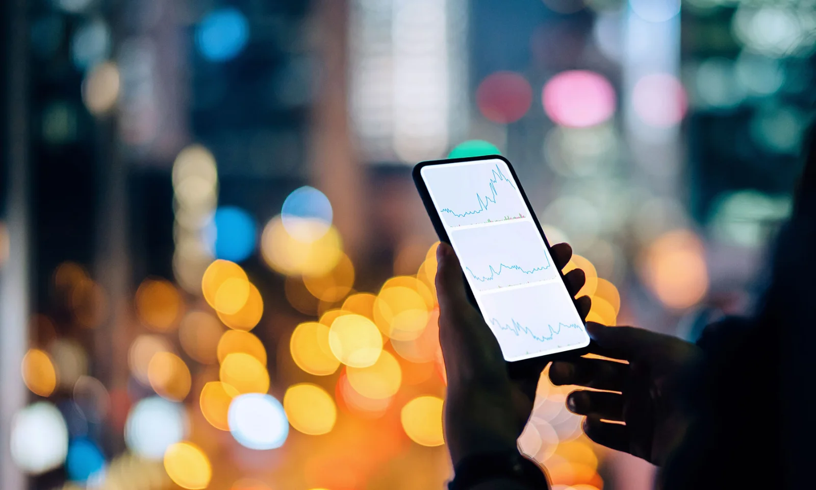 A person holding a smartphone displaying financial graphs, with a cityscape illuminated by vibrant lights in the background.