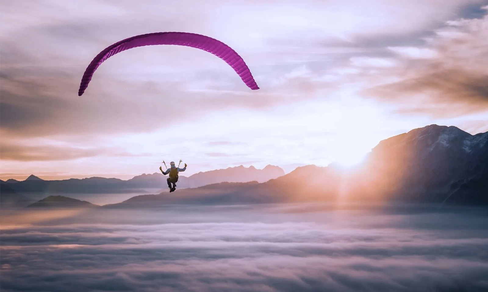 A paraglider silhouetted against a majestic sunrise, floating above a sea of clouds with mountain peaks in the background.