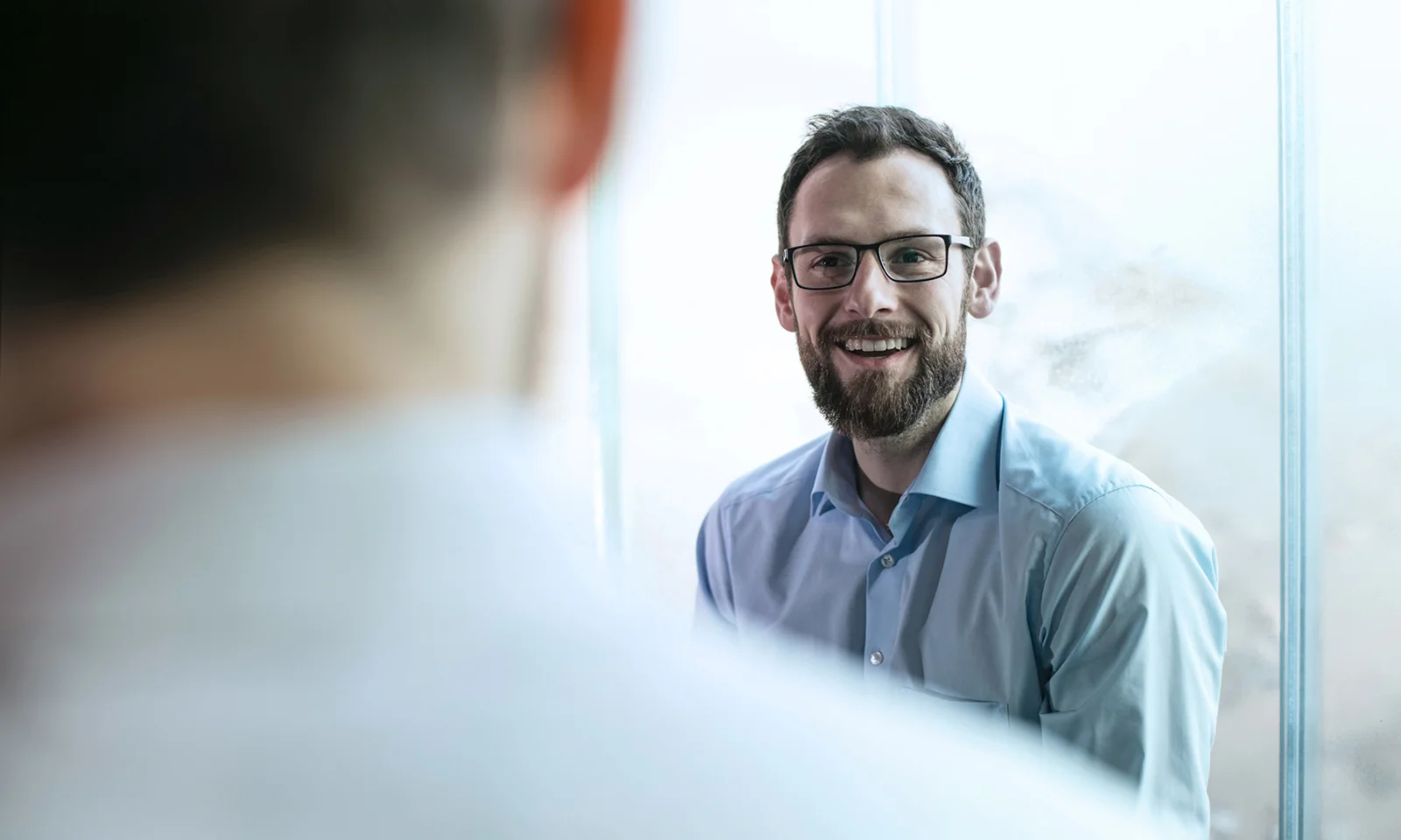 A business professional with glasses smiling during a conversation in a bright office setting