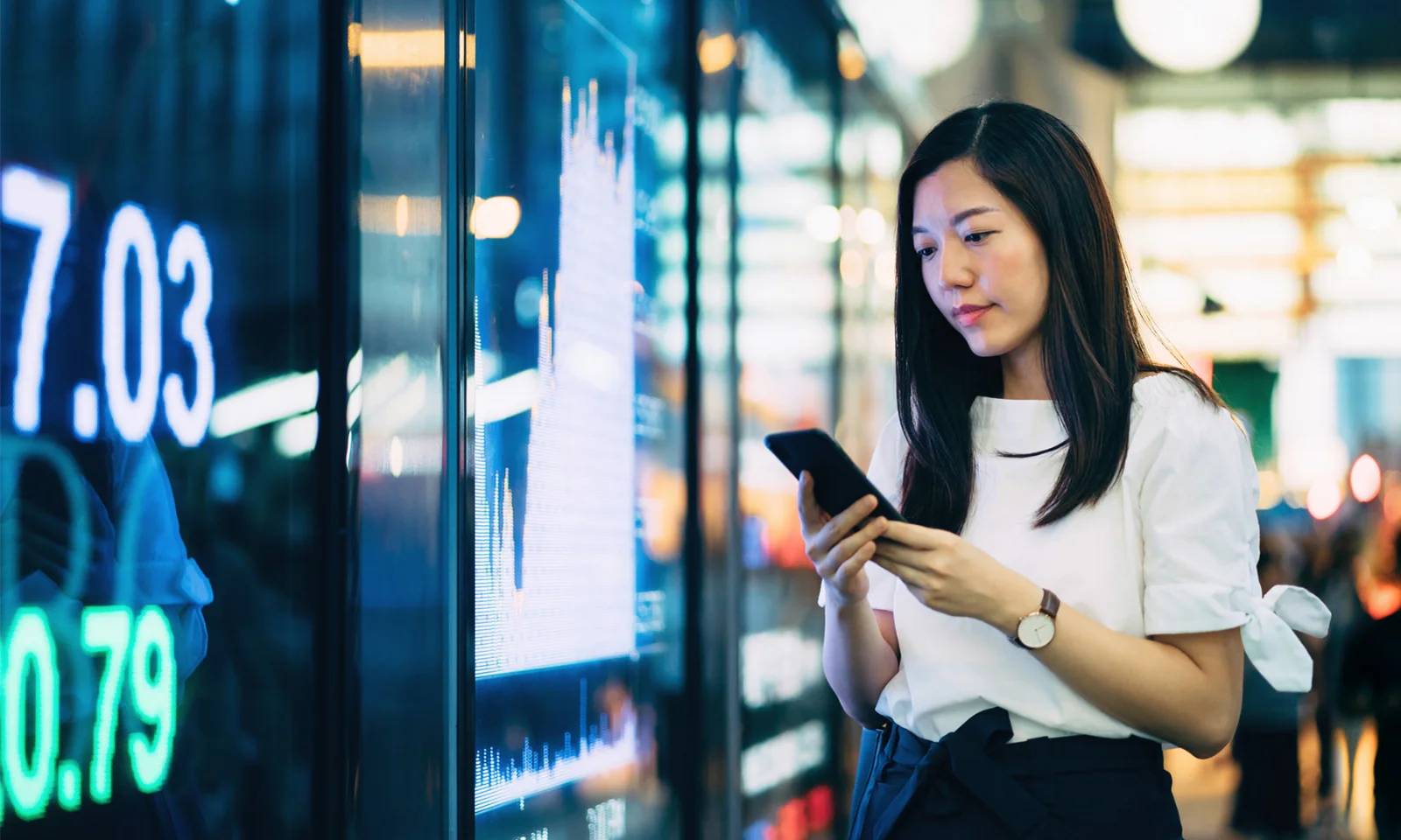 imagem mostra uma mulher usando seu smartphone enquanto está em pé na frente de um quadro eletrônico que exibe dados financeiros. Este visual simboliza a integração da sustentabilidade em tecnologia e práticas comerciais, destacando a importância do desenvolvimento sustentável na era digital moderna. O uso da tecnologia no monitoramento e promoção de iniciativas sustentáveis é um tema-chave representado nesta imagem.