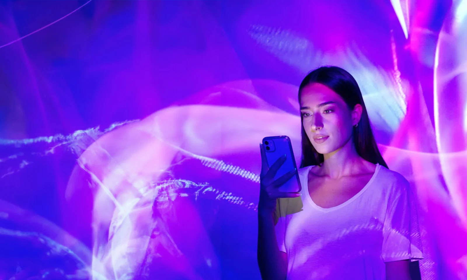 Una mujer que sostiene un teléfono inteligente se encuentra frente a una pantalla de luz vibrante y abstracta de color púrpura y rosa.