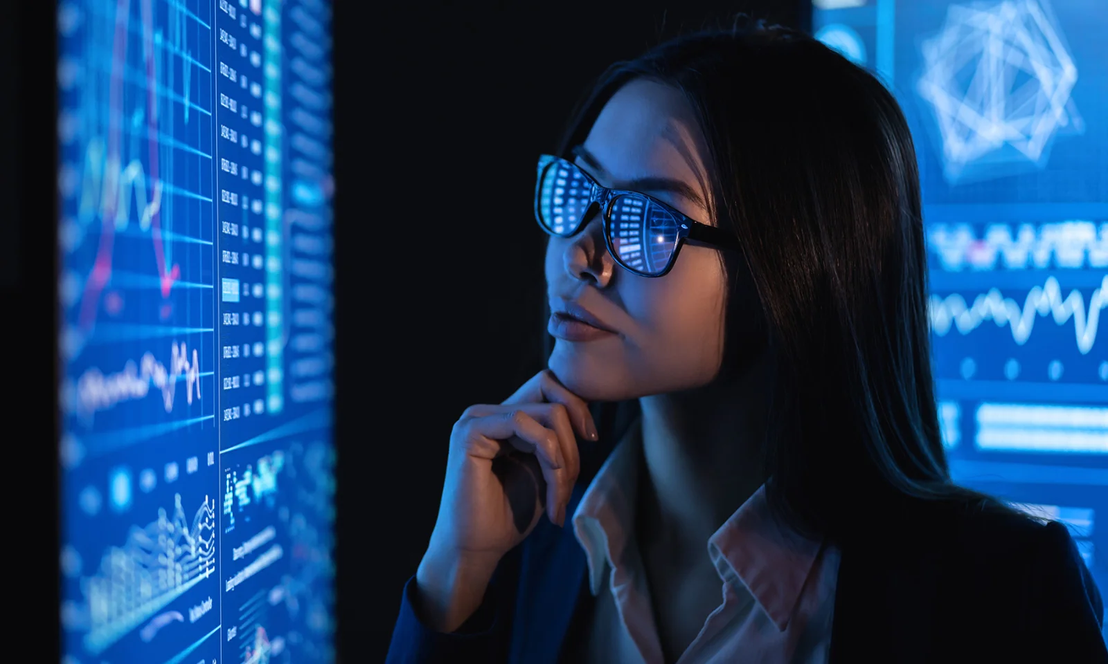 A woman in glasses, reflecting data charts, looks intently at multiple digital screens displaying various analytics and graphs.