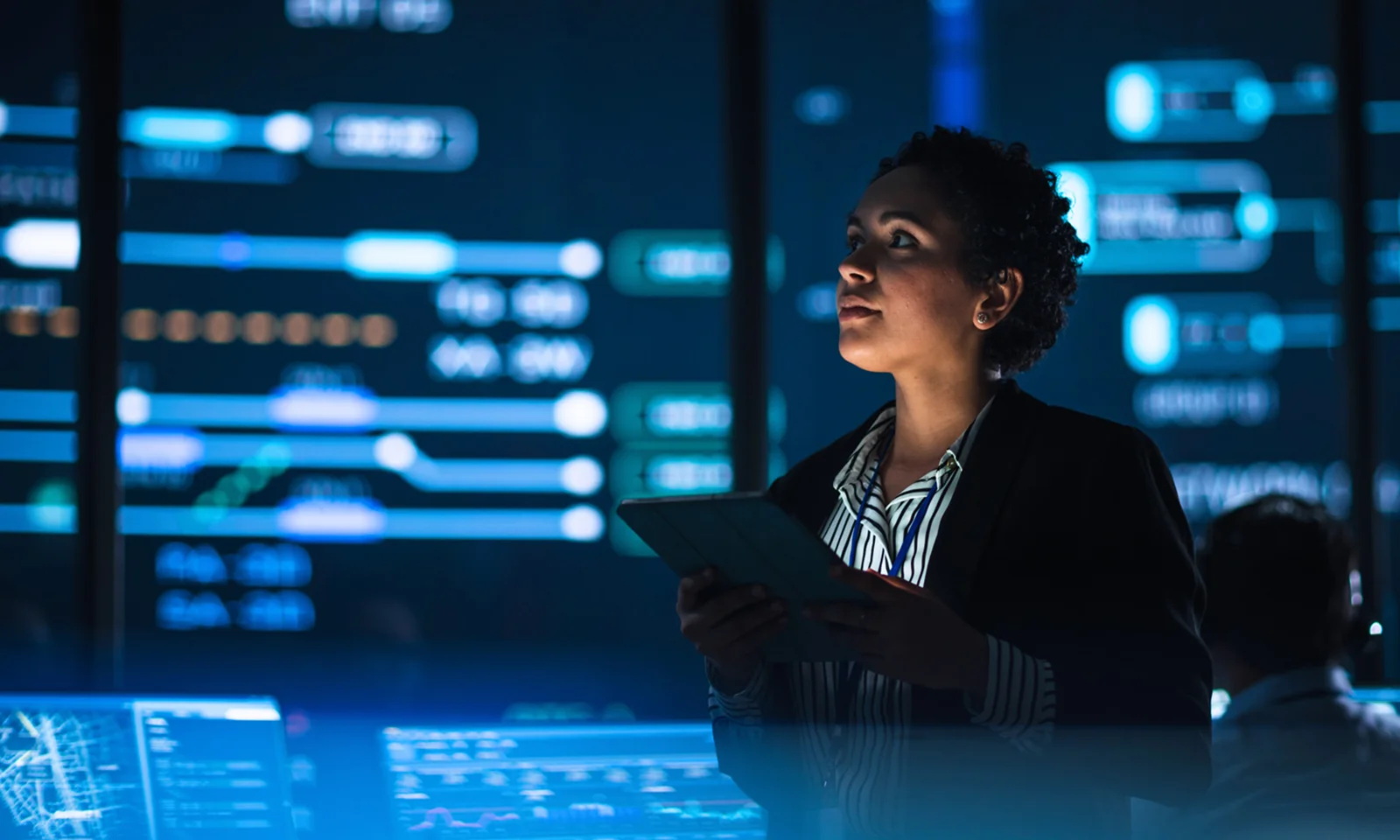 An IT professional using a tablet while overseeing digital operations in a high-tech control room with multiple data displays.