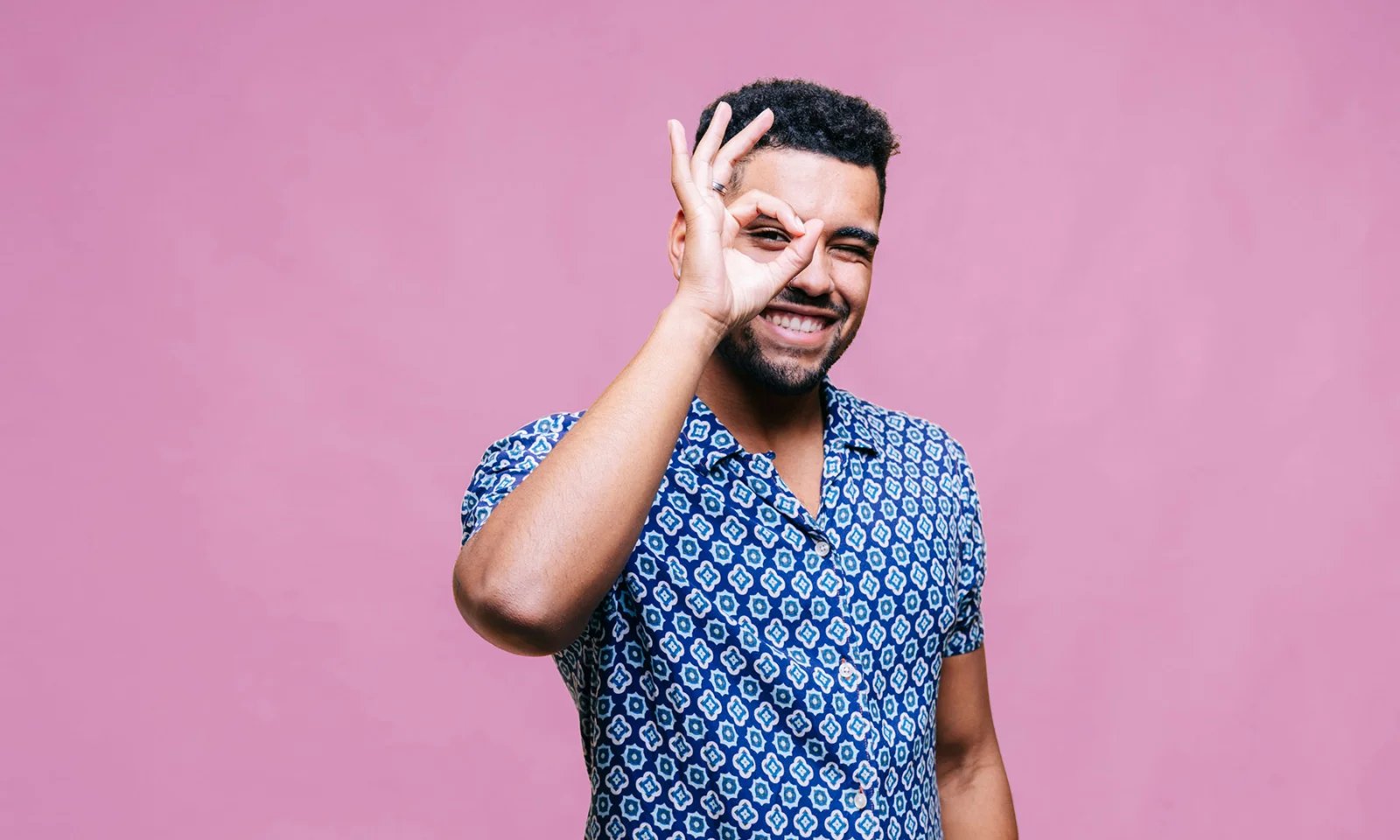 A cheerful young man making an &#039;OK&#039; sign with his hand against a pink background.