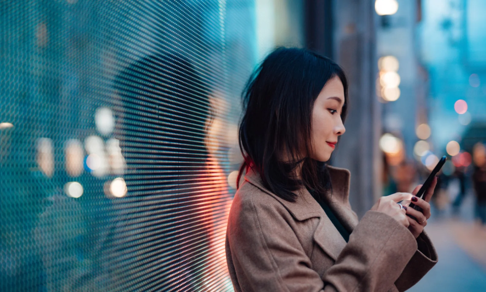 Eine Frau steht nachts vor einem digitalen Bildschirm und benutzt ihr Smartphone. Das Licht des Bildschirms spiegelt sich in ihrem Gesicht und der Umgebung und erzeugt eine moderne, technisch versierte Atmosphäre.