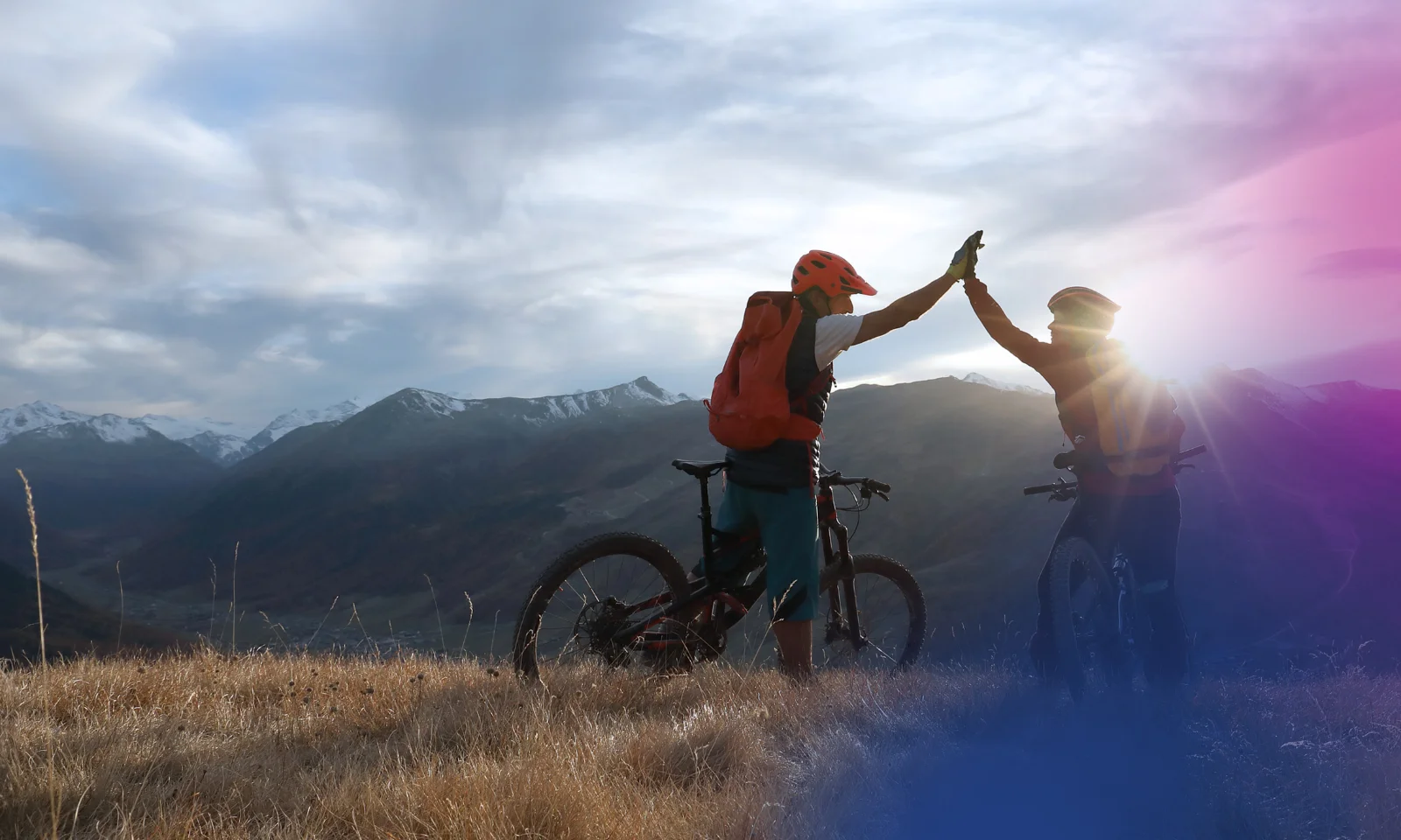 Two cyclists giving a high-five on a mountaintop, with a stunning landscape of mountains and valleys in the background, symbolising teamwork and shared achievement.