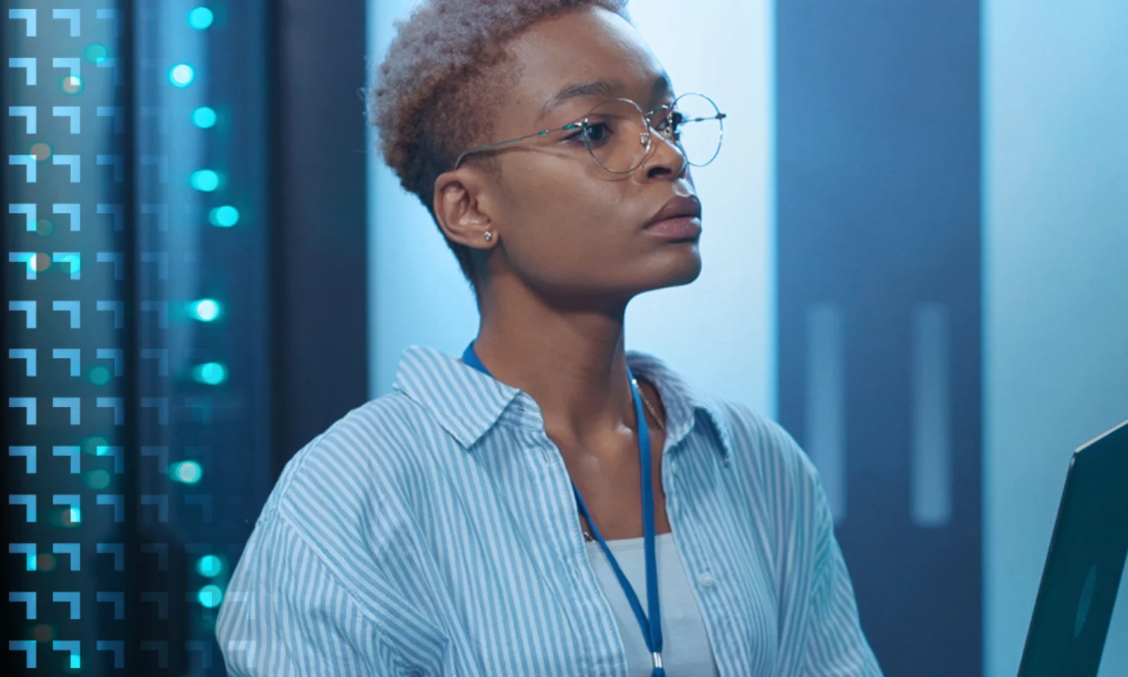 A focused tech professional with short hair and glasses analyzes data on a laptop in a blue-lit server room