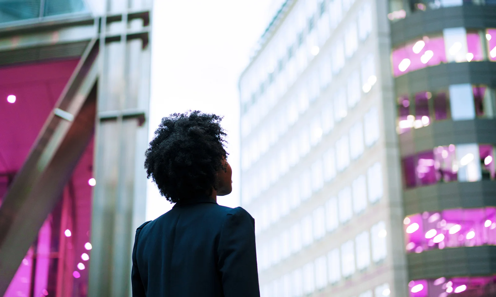 A person with natural hair, wearing a dark blazer, gazing up at modern office buildings with illuminated windows in a city environment.