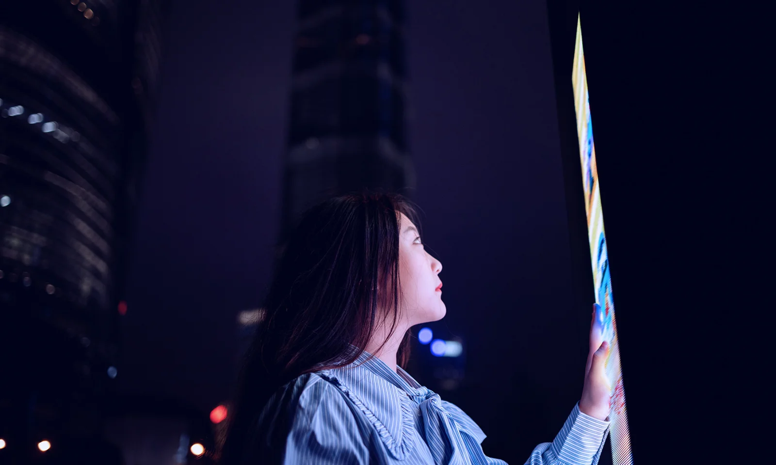 A woman interacts with a large, illuminated screen at night, set against the backdrop of a cityscape with towering buildings.